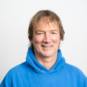 Foto: Bernd Drüke. Er hat helle Haut, braun-graue mittellange Haare und trägt einen blauen Pulli mit Kapuze. Er lächelt freundlich.