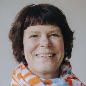 Foto von Ilona Hartmann. Sie lacht, trägt einen bunten Schal und hat helle Haut und dunkle mittellange Haare.