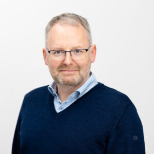 Foto von Dr. Jan Stewen. Er lächelt leicht und hat kurze graue Haare und helle Haut. Er trägt ein hellblaues Hemd mit dunkelblauem Pulli darüber und eine Brille.