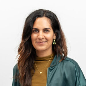 Foto: Jenny Heinz. Sie hat braune Haut, dunkle lange Haare und trägt einen braunen Pulli und eine grüne Jacke. Sie trägt Schmuck und lächelt freundlich.
