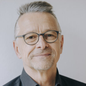 Foto von Jörg Gusek. Er lächelt, trägt eine Brille und hat graue Haare, die oben länger sind als an den Seiten. Er hat helle Haut.