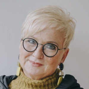 Foto von Dr. Margret Pohl. Sie lächelt und neigt den Kopf leicht. Sie hat kurze hellblonde Haare, trägt markanten Schmuck und eine Brille.