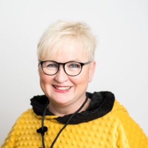 Foto von Dr. Margret Pohl. Sie lacht und hat hellblonde kurze Haare und helle Haut. Sie trägt eine schwarze Brille und einen gelb-schwarzen modischen Pulli.