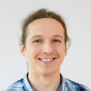 Foto von Martin Röthig. Er lacht freundlich und hat lange braune Haare, die zusammengebunden sind.