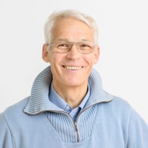 Foto von Dr. Michael Berkhoff. Er lächelt freundlich und hat kurze weiße Haare und helle Haut. Er trägt einen hellblauen Strickpulli und eine Brille.