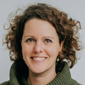 Foto von Nina Behringer. Sie lächelt freundlich, hat braune lockige Haare und helle Haut.