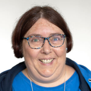 Foto von Sarah Borg. Sie lacht freundlich, hat helle Haut und trägt braune kurze Haare und eine gemusterte Brille.