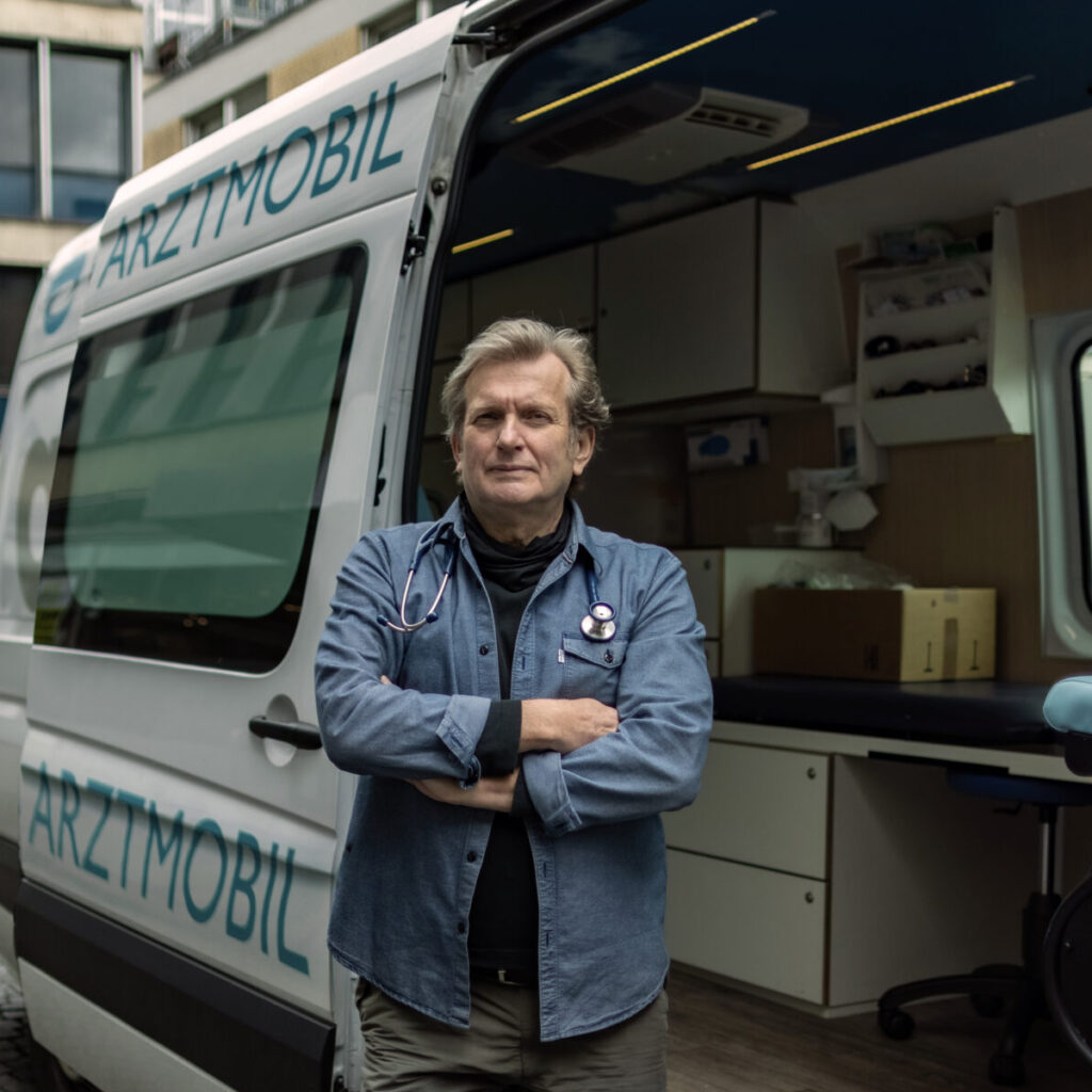 Foto von Prof. Dr. Gerhard Trabert mit verschränkten Armen vor seinem Arztmobil, 2021. Er hat graue kurze Haare, helle Haut und trägt ein Jeanshemd. Um seinen Hals hängt ein Stethoskop.