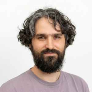 Foto: Johannes Lauxen. Er hat helle Haut, schwarz-graue Haare und einen schwarzen Bart. Er trägt ein lila Shirt und lächelt freundlich.